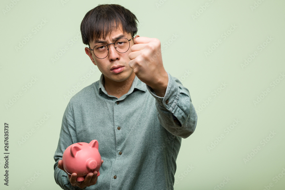 Young asian man holding a piggy bank showing fist to front, angry expression Young asian man holding a piggy bank showing fist to front, angry expression
