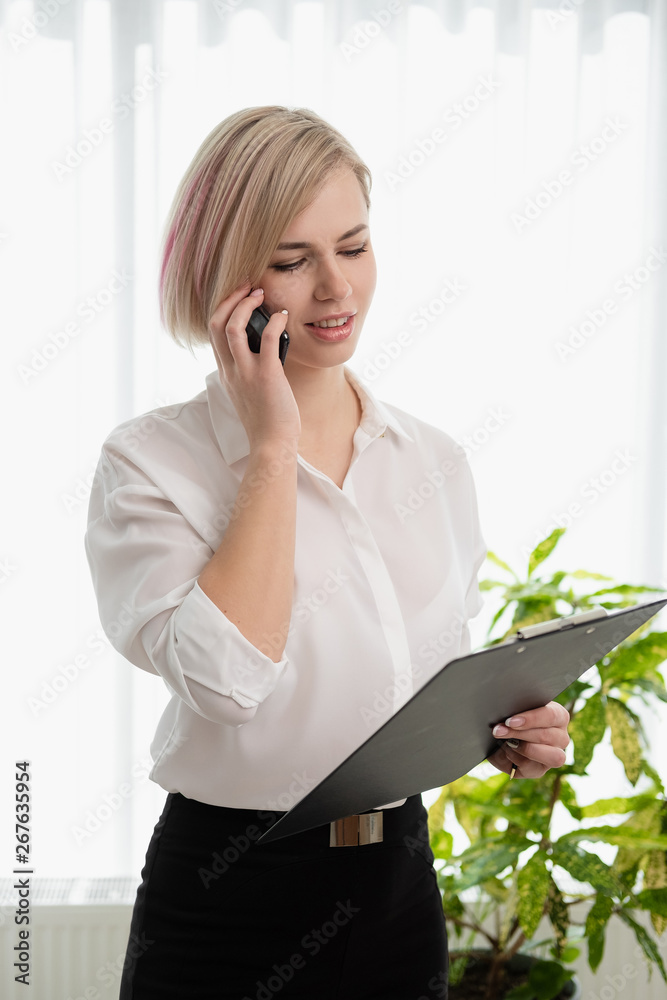 Young beautiful blonde girl with short hair in a white shirt is standing bright in the office against the window. Holds a notebook and smartphone. Talking on the phone. Young beautiful blonde girl with short hair in a white shirt is standing bright in the office against the window. Holds a notebook and smartphone. Talking on the phone.