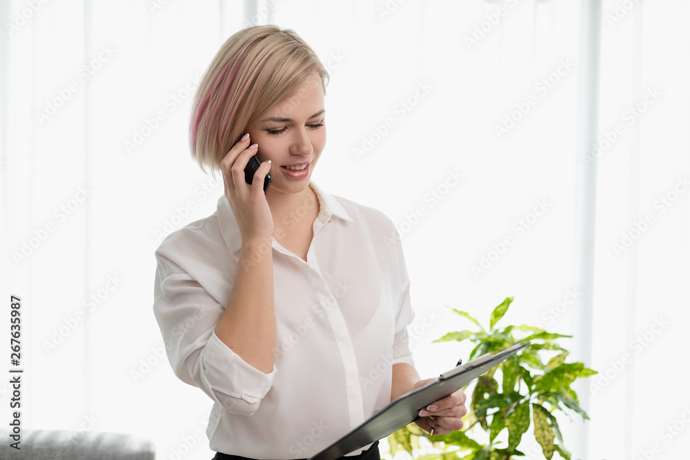 Young beautiful blonde girl with short hair in a white shirt is standing bright in the office against the window. Holds a notebook and smartphone. Talking on the phone. Young beautiful blonde girl with short hair in a white shirt is standing bright in the office against the window. Holds a notebook and smartphone. Talking on the phone.