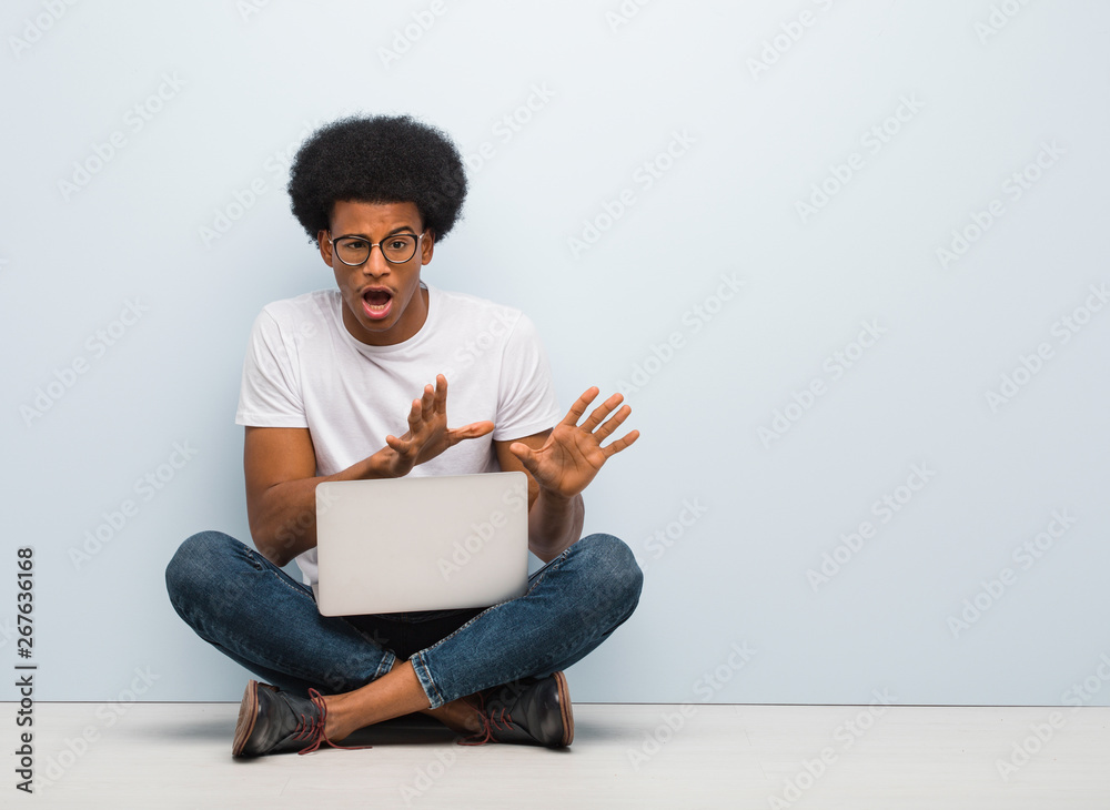 Young black man sitting on the floor with a laptop rejecting something doing a gesture of disgust Young black man sitting on the floor with a laptop rejecting something doing a gesture of disgust
