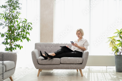Young beautiful blonde girl with short hair in a white shirt is sitting on the sofa in bright in the office against the window. Holds a notebook and smartphone. Talking on the phone. © spaskov