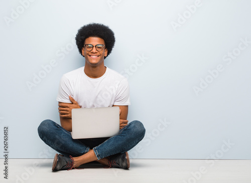 Young black man sitting on the floor with a laptop crossing arms, smiling and relaxed
