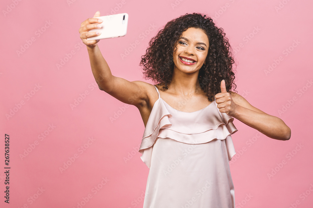 Portrait of a pretty young afro american woman in dress while standing and taking a selfie isolated over pink background. Portrait of a pretty young afro american woman in dress while standing and taking a selfie isolated over pink background.