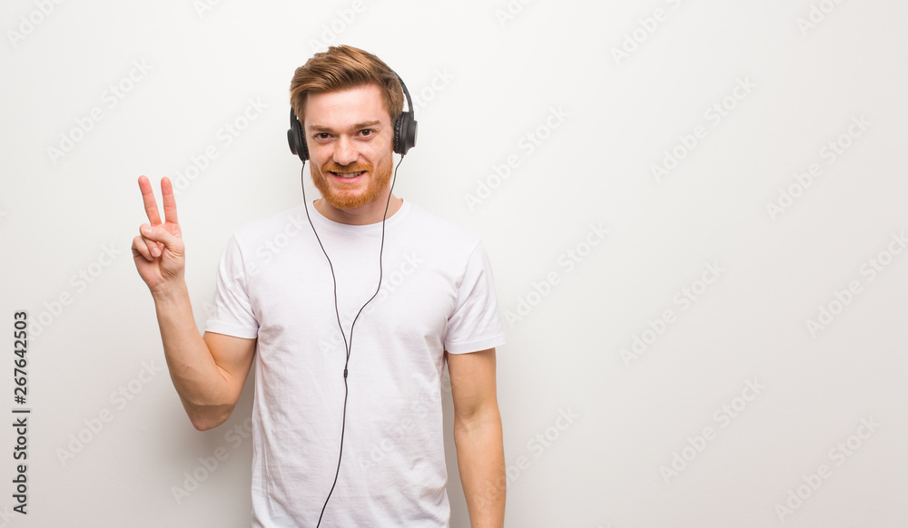 Young redhead man showing number two. Listening to music with headphones.