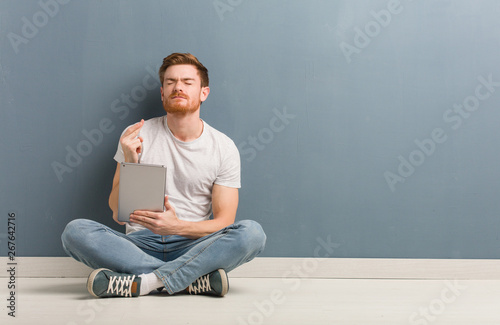 Young redhead student man sitting on the floor crossing fingers for having luck. He is holding a tablet.