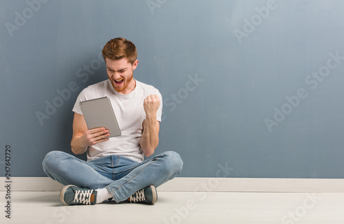 Young redhead student man sitting on the floor surprised and shocked. He is holding a tablet.