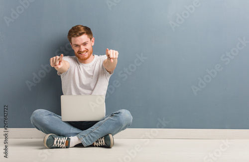 Young redhead student man sitting on the floor cheerful and smiling pointing to front. He is holding a laptop.