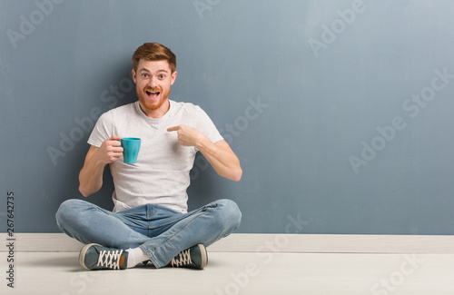 Young redhead student man sitting on the floor surprised, feels successful and prosperous. He is holding a coffee mug.