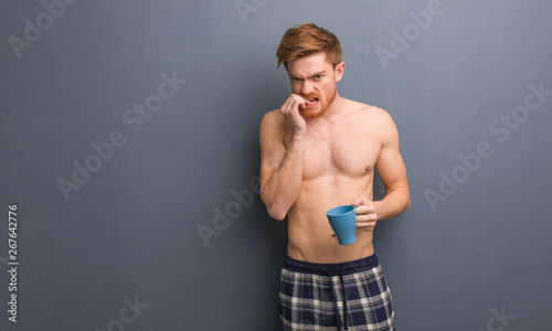 Young shirtless redhead man biting nails, nervous and very anxious. He is holding a coffee mug.