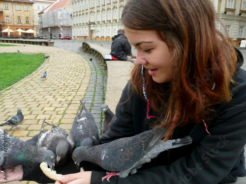 Young white girl feeding wild pigeons with bread in the city park on a cold day. Brick pavement and old architecture in the background.