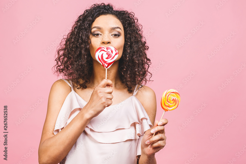 Happy sexy american afro girl eating lollipop. Beauty glamour model woman holding pink sweet colorful lollipop candy, isolated on pink background. Sweets.