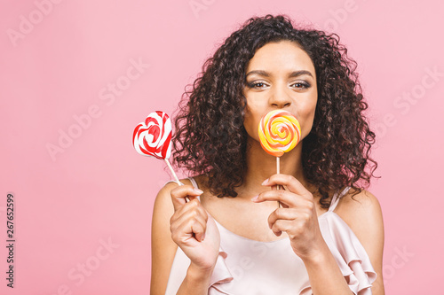 Happy sexy american afro girl eating lollipop. Beauty glamour model woman holding pink sweet colorful lollipop candy, isolated on pink background. Sweets.