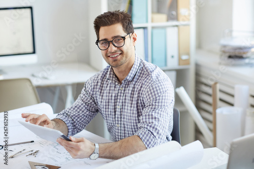 Portrait of young engineer smiling at camera while using digital tablet sitting at desk in office, copy space photo