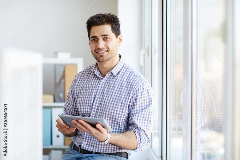 Portrait of handsome Middle-Eastern man looking at camera while posing by window holding tablet, copy space