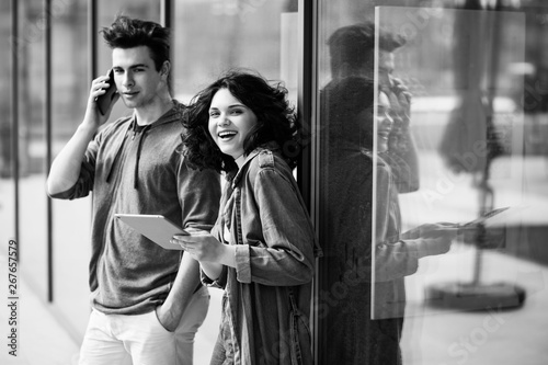 Young cute couple - a boy and a girl standing near a glass building. The couple spend time together. The guy speaks on the phone, and the girl holds a tablet in her hands