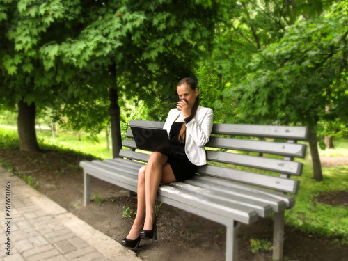 Young woman in a business suit drinks coffee and works on a laptop while sitting on a bench in the park, side view. Lonely adult girl works on the Internet enjoying nature outdoors