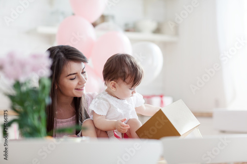 Beautiful young mother smiling while her baby daughter opening birthday gift box. Room decorated with flowers and balloons