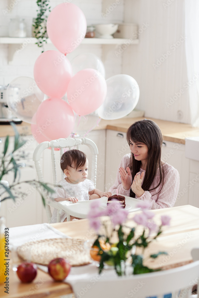 Loving young mother clapping hands while baby daughter blowing out candle on her first birthday cake sitting in highchair