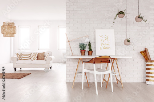 Open space white flat interior in real photo with desk with poster, lamp and fresh plants on brick wall and couch with pillows in the background