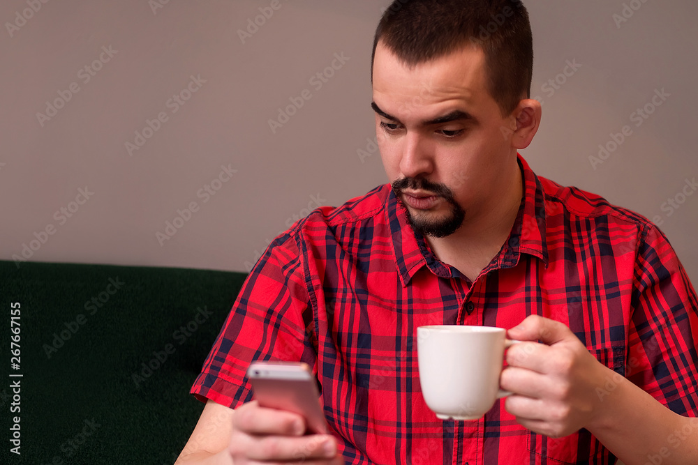 Middle-aged man with surprised face sitting on green sofa with mug of coffee and reading or watching something on smartphone Middle-aged man with surprised face sitting on green sofa with mug of coffee and reading or watching something on smartphone