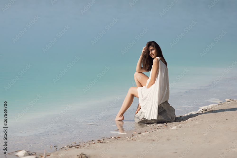 Young very beautiful girl with long hair in a white dress by the lake. The girl sits on a stone and enjoys the rest. Young very beautiful girl with long hair in a white dress by the lake. The girl sits on a stone and enjoys the rest.
