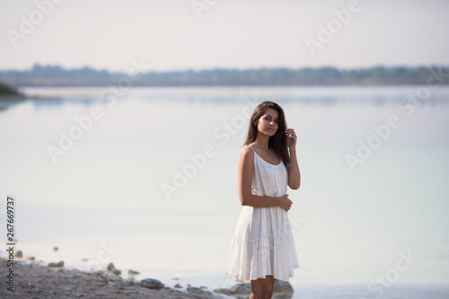 Young very beautiful girl with long hair in a white dress by the lake. The girl is enjoying the rest.