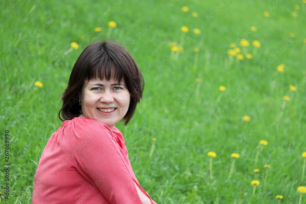Happy middle aged woman in pink dress sitting on a green meadow with dandelion flowers. Concept of happiness, positive emotions, joy, ecology, leisure outdoors Happy middle aged woman in pink dress sitting on a green meadow with dandelion flowers. Concept of happiness, positive emotions, joy, ecology, leisure outdoors