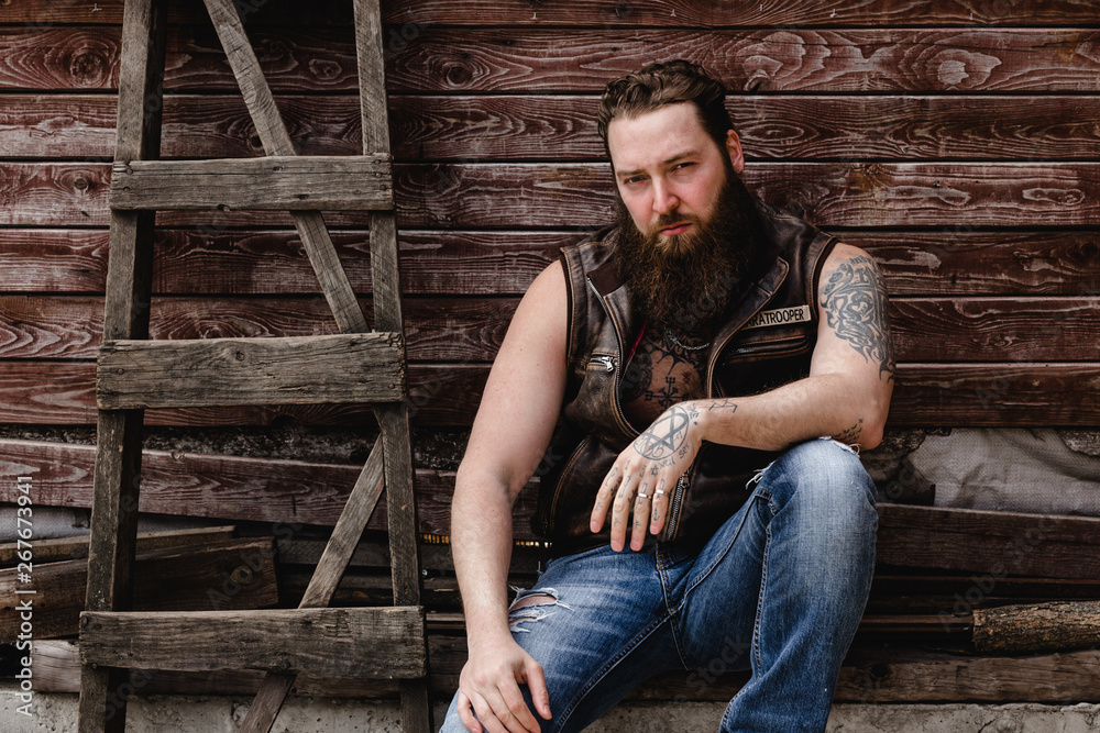 Strong brutal man with a beard and tattoos on his hands dressed in leather vest and jeans  sits on a wooden wall background next to a wooden ladder outside