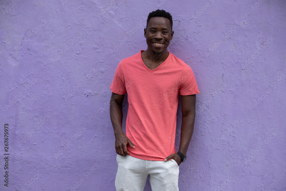Smiling African American man model posing in empty living coral color t-shirt standing against violet wall background