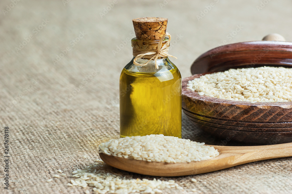 Glass bottle of sesame oil and raw sesame seeds in wooden shovel or spoon and in bowl on burlap sack. Uncooked sesame background concept with copy space Glass bottle of sesame oil and raw sesame seeds in wooden shovel or spoon and in bowl on burlap sack. Uncooked sesame background concept with copy space