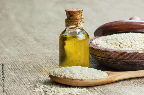 Glass bottle of sesame oil and raw sesame seeds in wooden shovel or spoon and in bowl on burlap sack. Uncooked sesame background concept with copy space