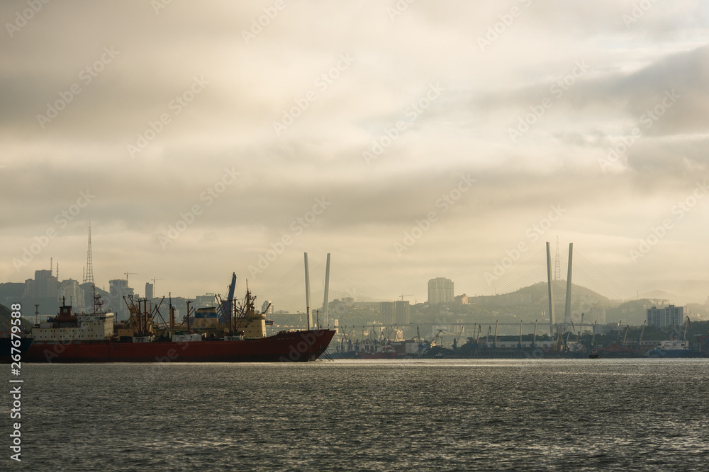 The sea trade port of Vladivostok on the background of the Golden Bridge across the Golden Horn Bay in the rays of the rising sun