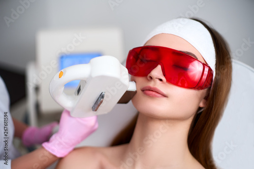 Beautician doctor doing laser RF rejuvenation for pretty young woman face at beauty salon. Elos epilation hair removal procedure. Aesthetic facial acne treatment skin care. Hardware ipl cosmetology