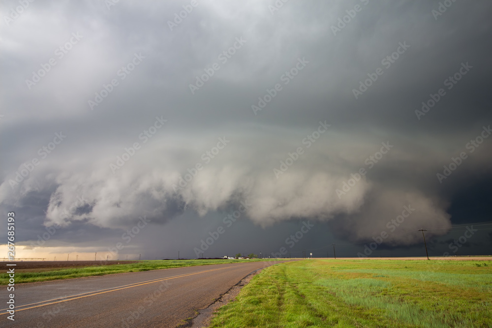 A big supercell storm with a shelf cloud and a wall cloud looms over a road in the rural countryside.
