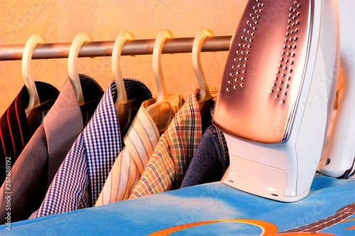 Colored shirts on the hanger, electric iron, Ironing Board in the Laundry room. Ironing after washing, heated by the plane of the sole of the iron, the fabric acquires a smooth surface.  photo