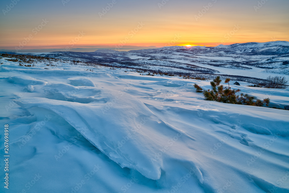 Landscape with snow drifts on the hillside. Sastrugi (zastrugi) formed on the surface of snow by wind erosion. View of the valley among the mountains. Beautiful northern sunset. Magadan Region, Russia