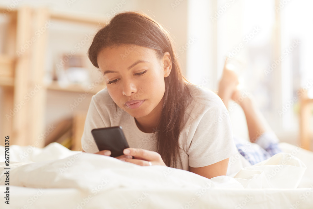 Portrait of beautiful Mixed-Race woman using smartphone lying on bed lit by sunlight, copy space Portrait of beautiful Mixed-Race woman using smartphone lying on bed lit by sunlight, copy space