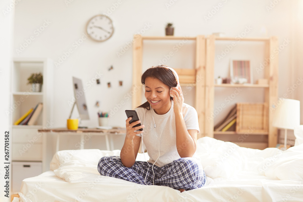 Portrait of smiling Mixed-Race woman using smartphone sitting on bed lit by sunlight, copy space Portrait of smiling Mixed-Race woman using smartphone sitting on bed lit by sunlight, copy space