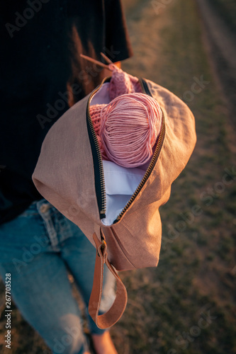 Close-up of a young woman holding a handmade bag with accessories for knitting: knitting needles, crochet hooks, pink ball of yarn, against a background of a field