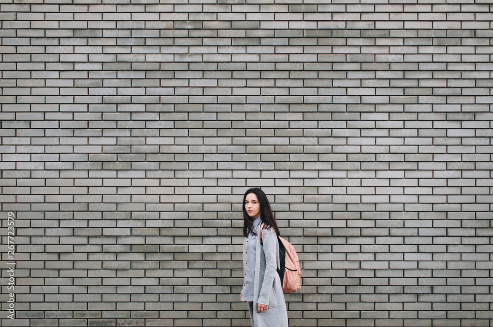 Beautiful attractive young woman with a backpack standing half a turn near the gray brick wall. Lonely look. Pensive girl. Gray colour. Copy space. Beautiful attractive young woman with a backpack standing half a turn near the gray brick wall. Lonely look. Pensive girl. Gray colour. Copy space.