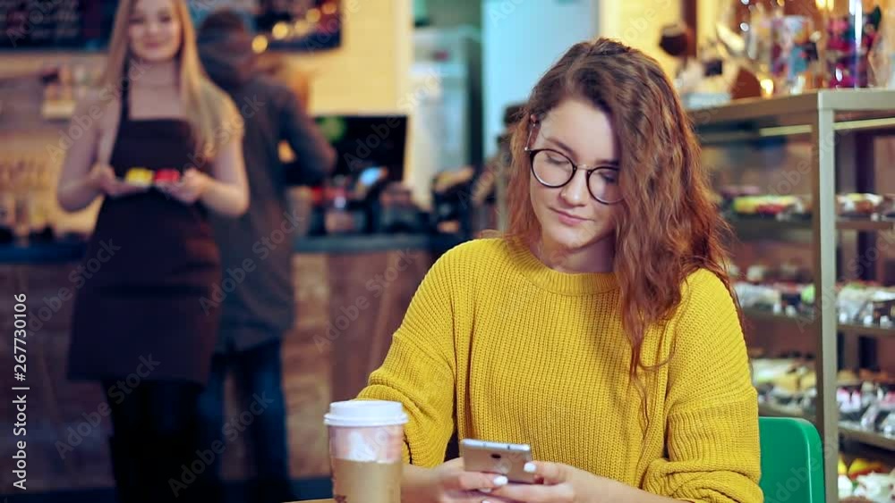 Beautiful girl in glasses sits at a table in a coffee shop, she looks into the smartphone. The waiter comes from a defocused background with dessert Beautiful girl in glasses sits at a table in a coffee shop, she looks into the smartphone. The waiter comes from a defocused background with dessert