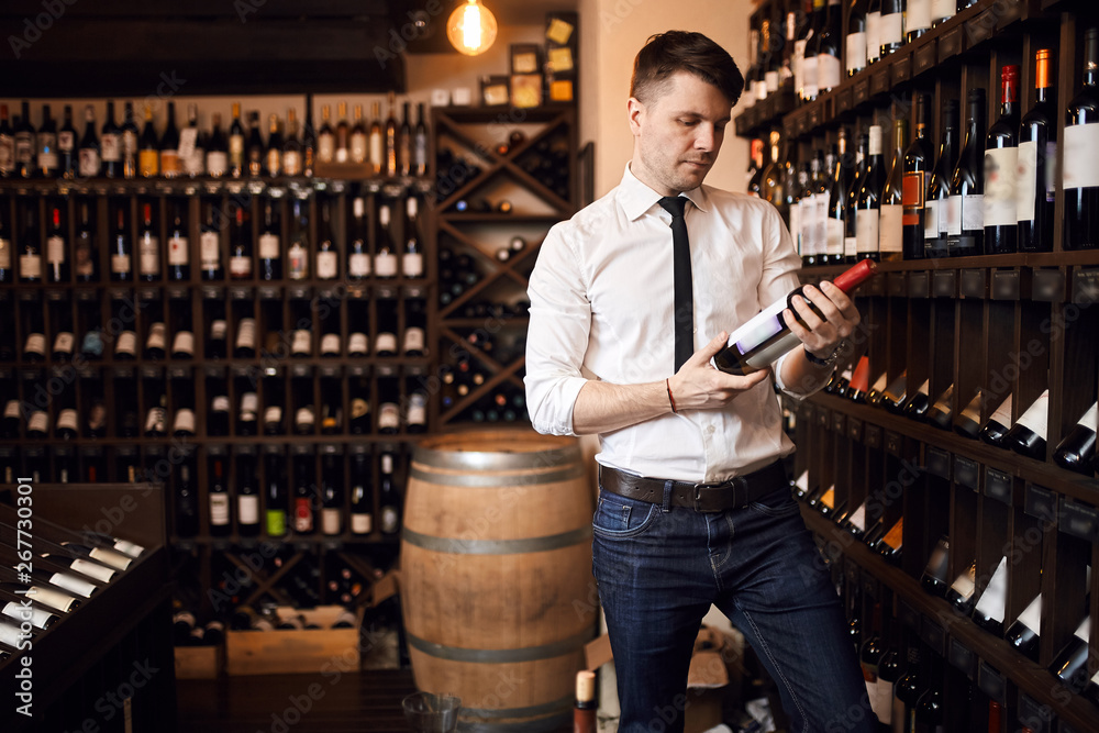 attractive sommelier wearing white shirt and trousers standing in wine store holding bottle of red wine , wine shelves on the background. copy space, interest