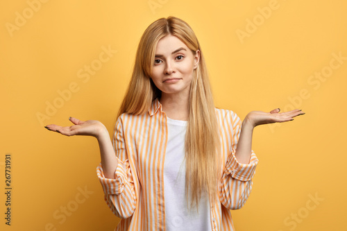 Puzzled hesitant girl with long fair hair dressed casually, shrugs shoulders as doesn t know answer, to be or not to be.isolated yellow background photo