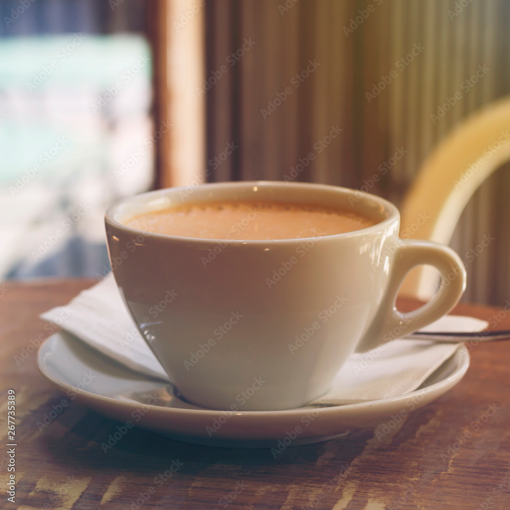 Fototapeta premium Cup of cappuccino coffee or orange thai tea on the brown wooden table in street cafe with sunlight highlight, toned. Close-up, copy space, square format
