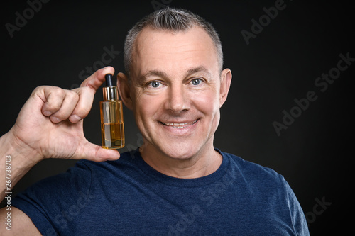 Handsome middle-aged man with bottle of serum for skin care on dark background