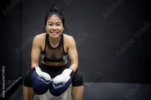 Confident Asian happy girl sitting and posing with blue boxing gloves on the chair nearby black wall in fitness gym. Healthy lifestyle with martial arts concept.