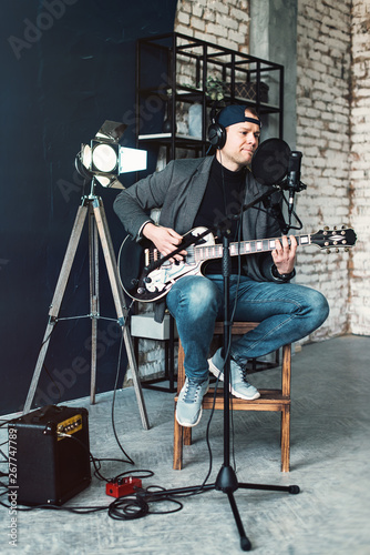 Close up of a man singer sitting on a stool in a headphones with a guitar recording a track in a home studio