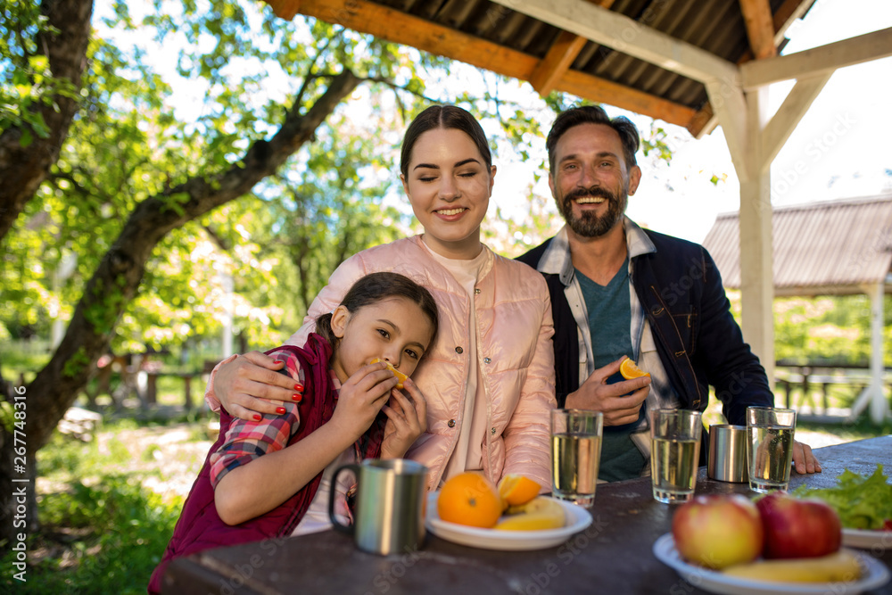 Happy Family Are Sitting By The Table In The Park. Plates With Different Fruits And Glasses With Water Are On The Table. Happy Family Are Sitting By The Table In The Park. Plates With Different Fruits And Glasses With Water Are On The Table.