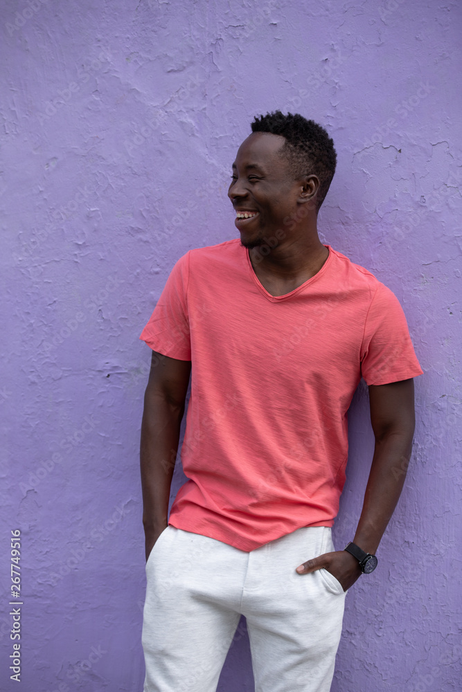 Smiling laughing African American man model posing in empty living coral color t-shirt standing against violet wall background Smiling laughing African American man model posing in empty living coral color t-shirt standing against violet wall background