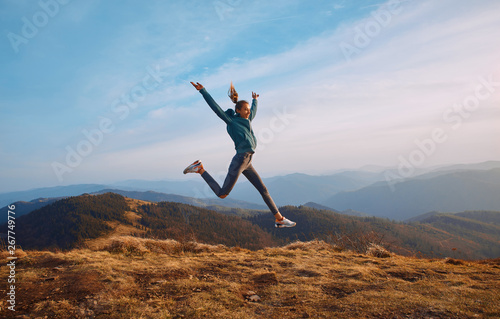 Happy woman hiker jumping on mountain ridge on blue cloudy sky and mountains background. Travel and active lifestyle concept.